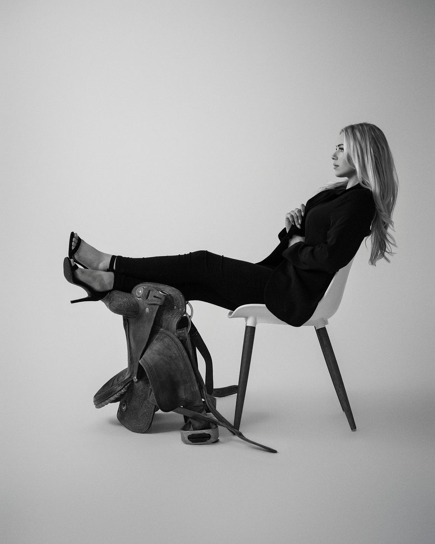 Black-and-white studio portrait of the founder seated in profile on a white chair, heels resting on a western saddle
