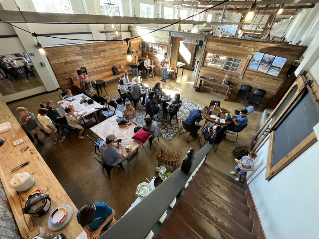 People seated around wooden tables under string lights in a warm, rustic room.