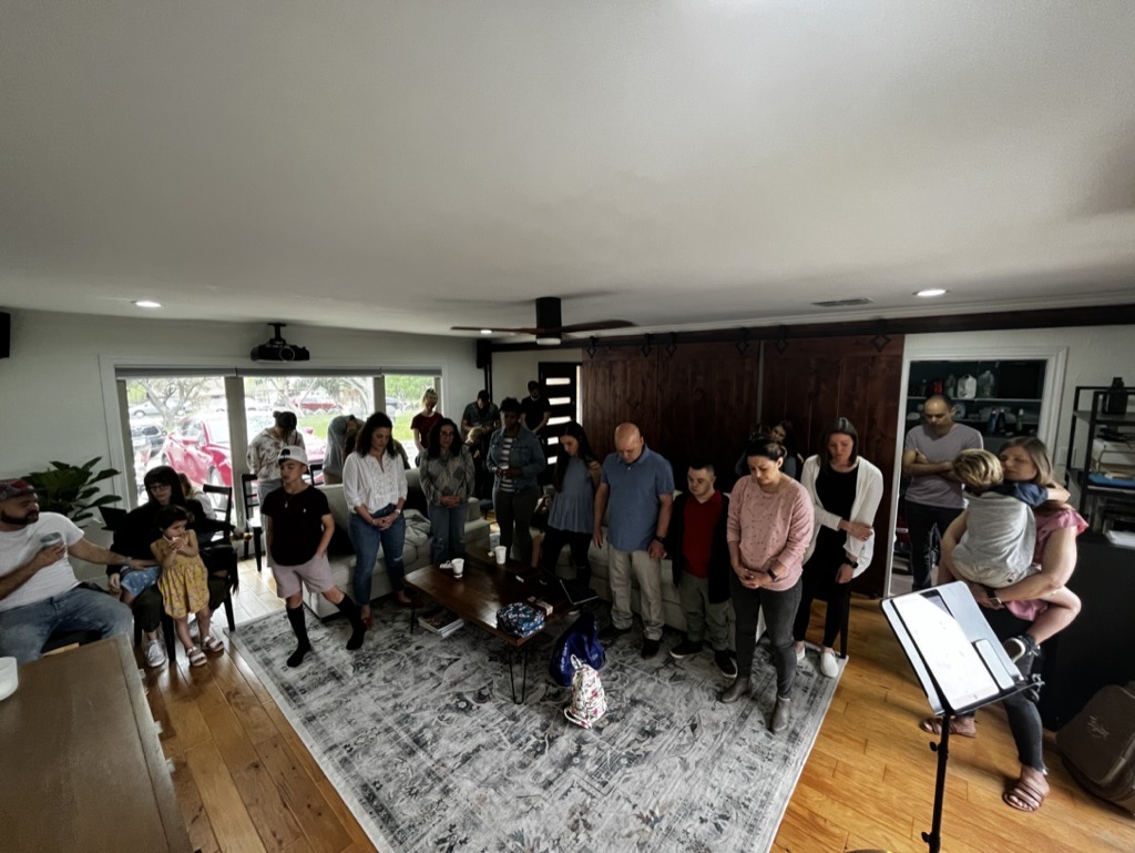 A crowded living room of people standing together, some with heads bowed in prayer.