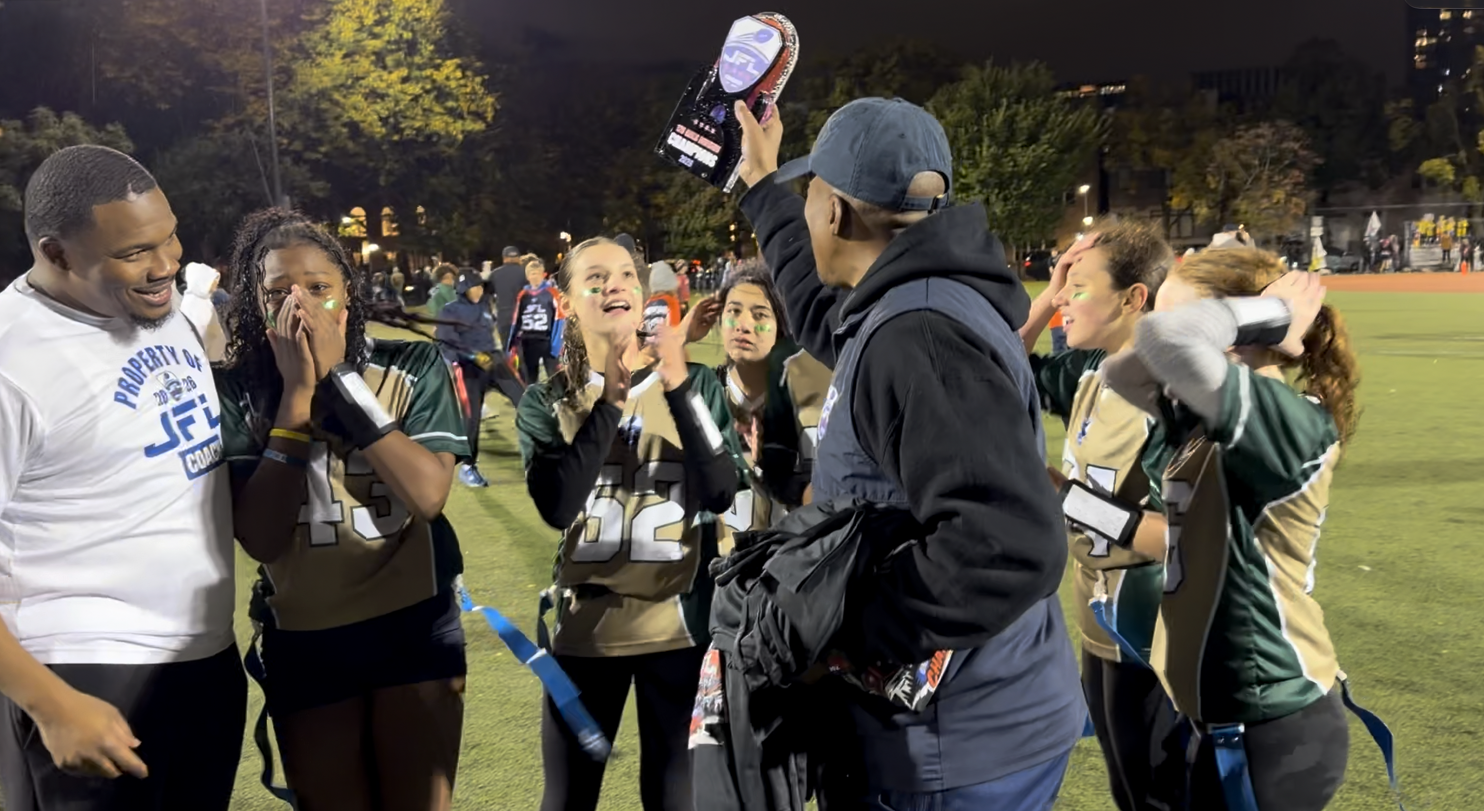 JFL champions celebrating with the 2026 championship trophy on the field at night