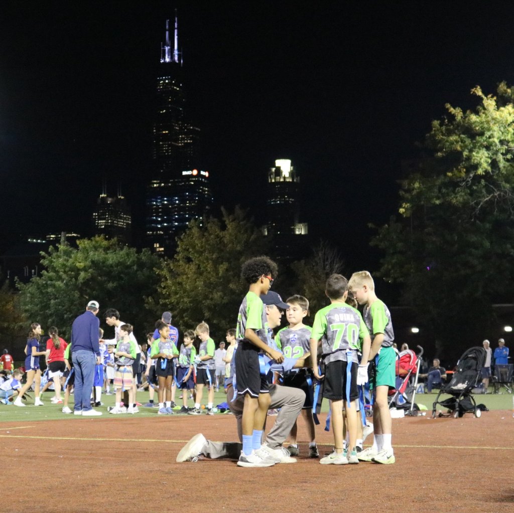 JFL coach with young players on the field at night with Chicago skyline in the background