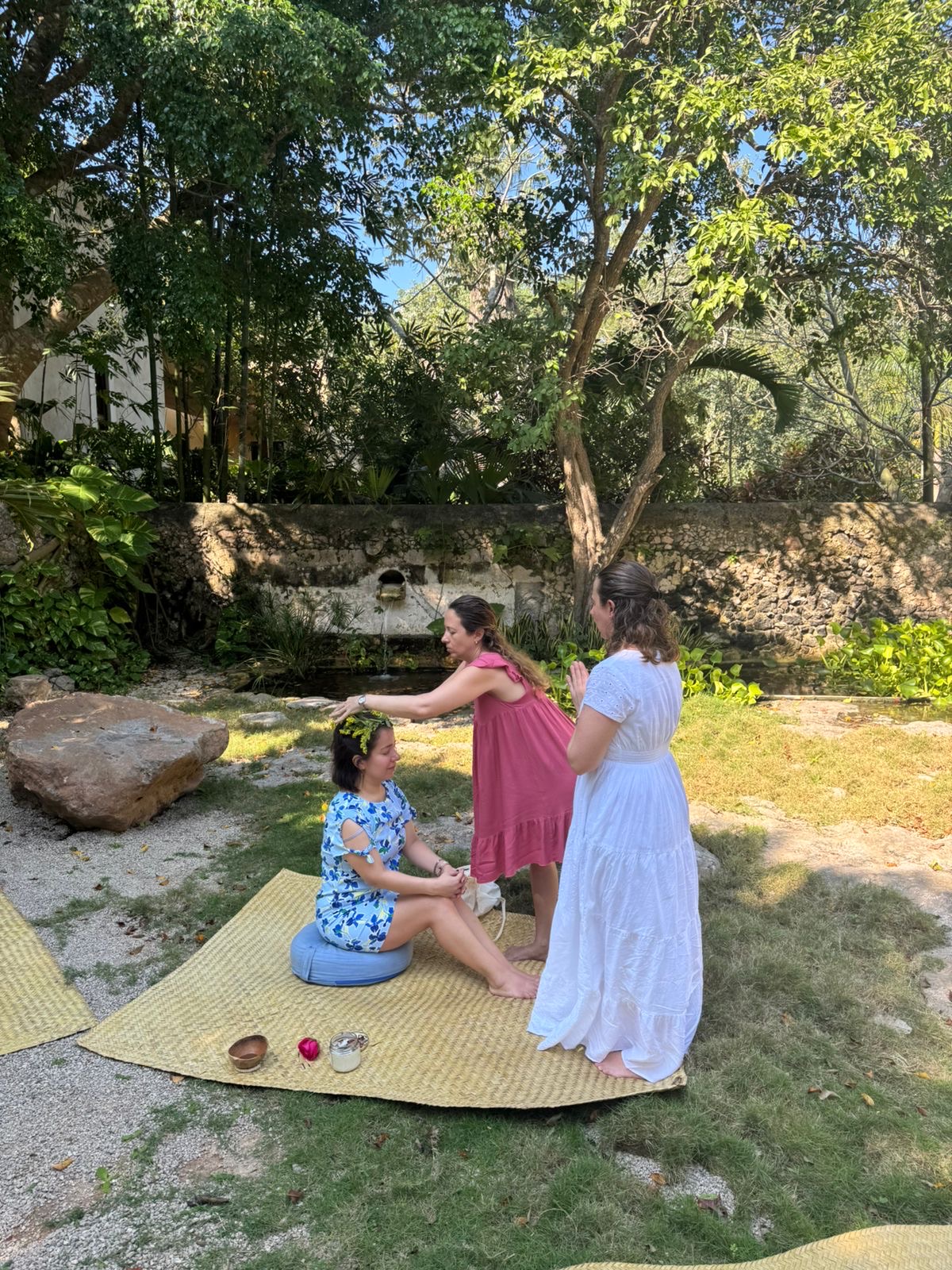 Tres mujeres realizando un rito de paso al aire libre en un jardín; una mujer coloca una corona de flores naturales a otra sentada sobre un petate