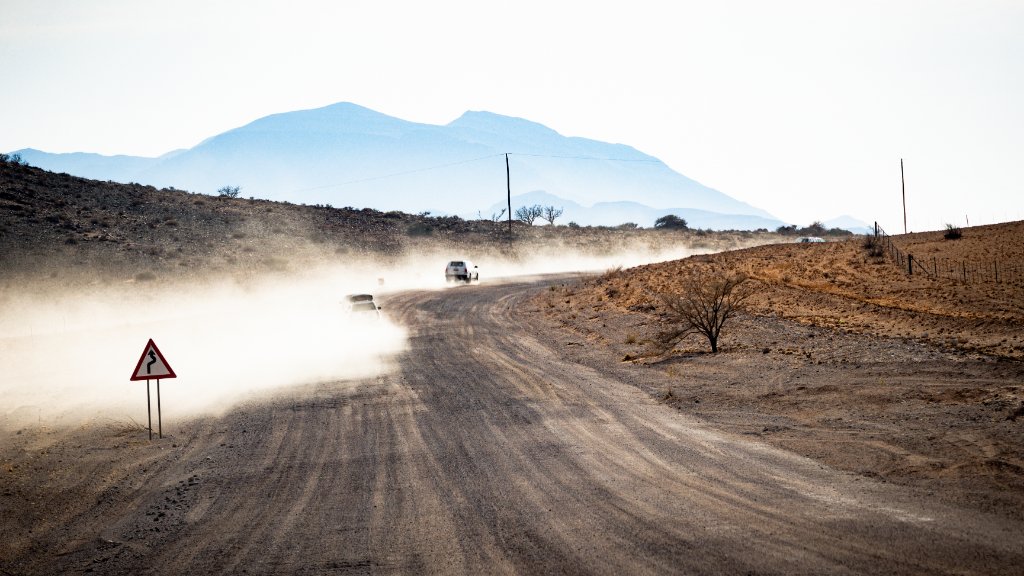 Two vehicles raising dust on a Namibian gravel road, mountains in the distance — a reminder that gravel drives take longer than the map says.