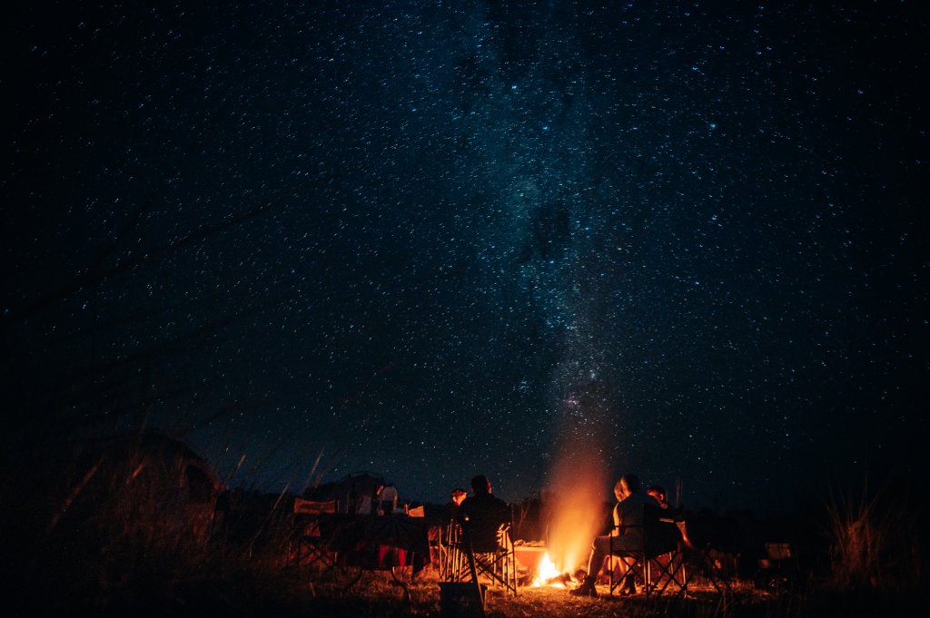 A brilliant, star-filled Milky Way arches over a desert camp and glowing campfire in Namibia, far from any light pollution.
