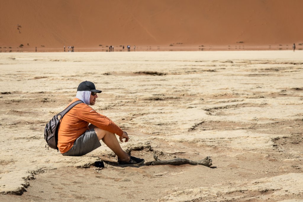 A lone traveller sits on the pale, cracked clay floor of Sossusvlei, dwarfed by a towering orange dune with tiny figures at its base and a weathered dead branch resting on the ground nearby.