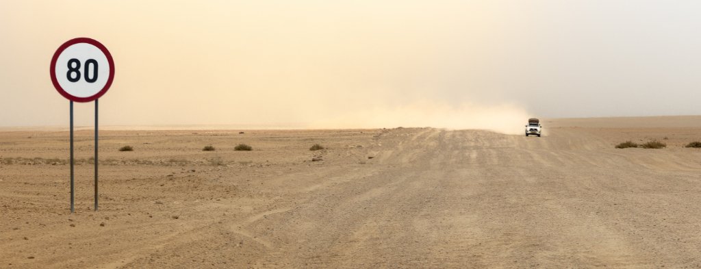 A lone gravel road runs dead straight into the empty Namibian distance, an 80 km/h sign at the roadside and a faint dust cloud on the horizon.