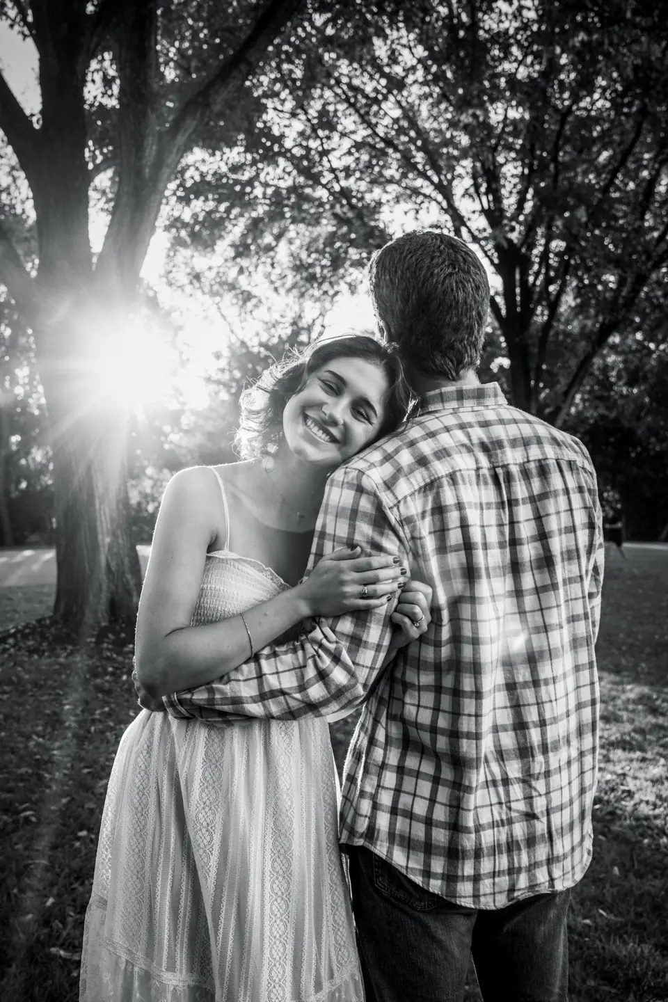 Black and white engagement photo of a couple hugging under a tree with sunlight behind them