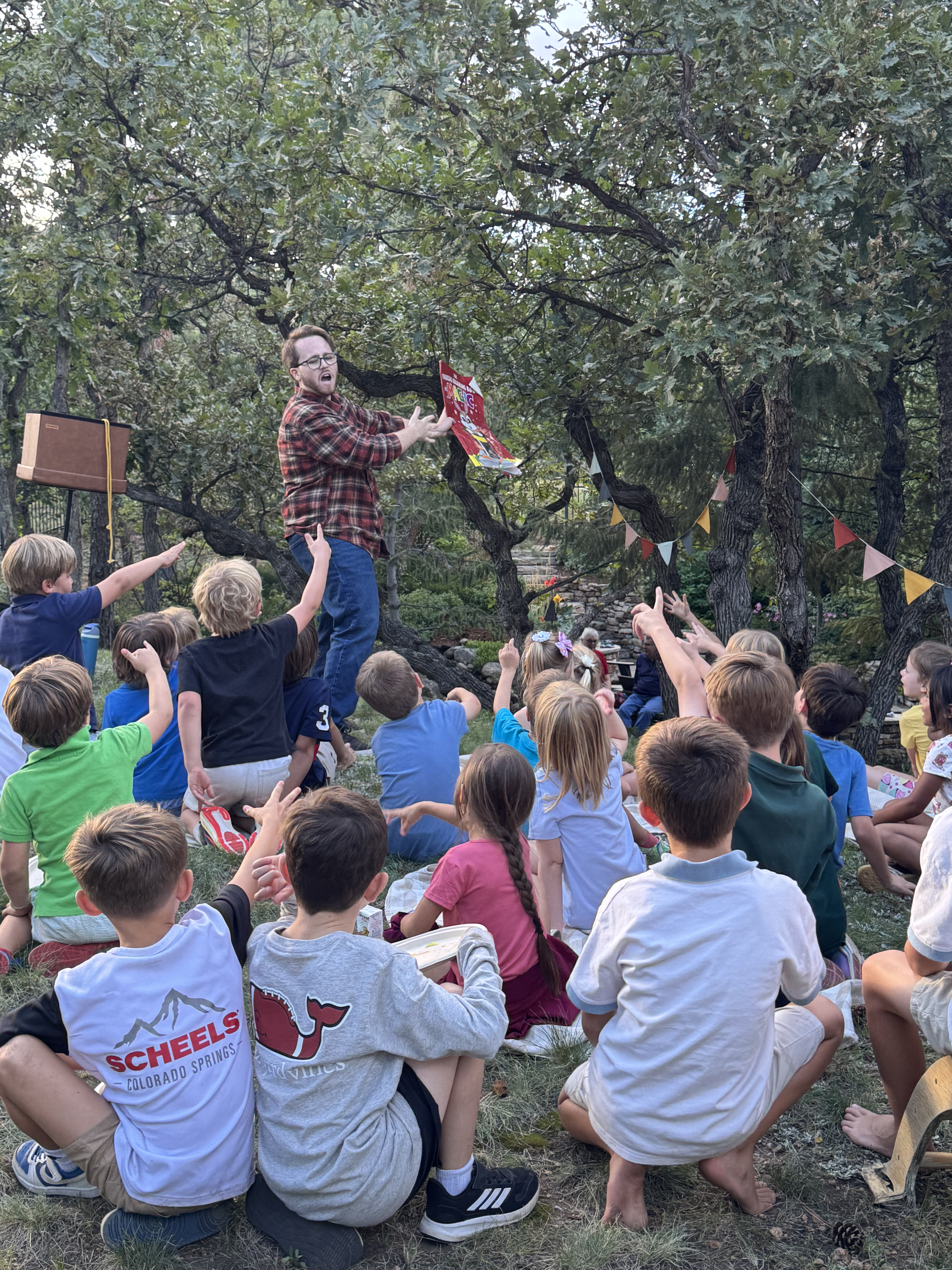 Kids raising their hands during a Braden Carlisle birthday party magic show in a backyard under oak trees