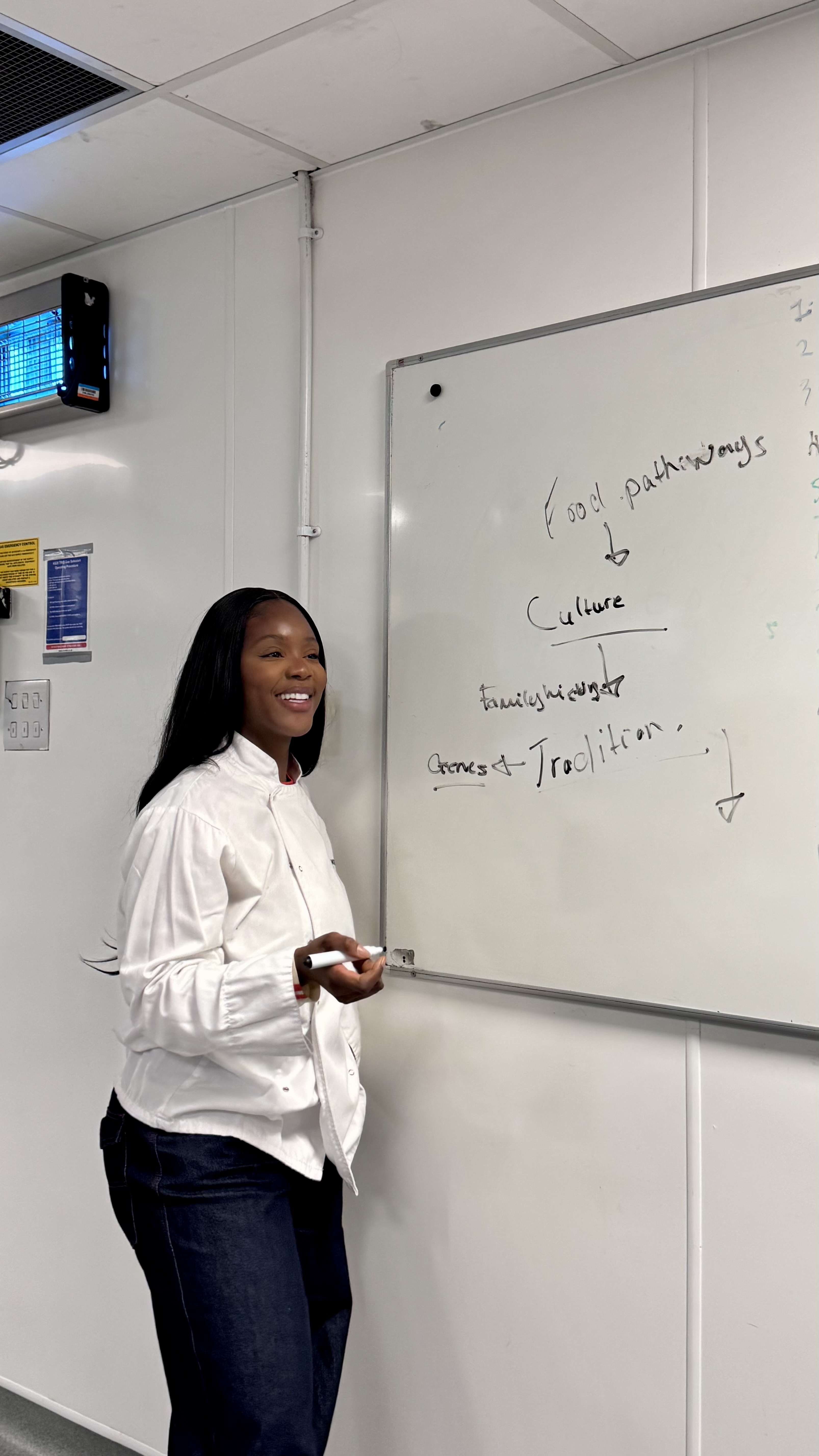 Chef Mona in her white chef jacket beside a food-pathways whiteboard