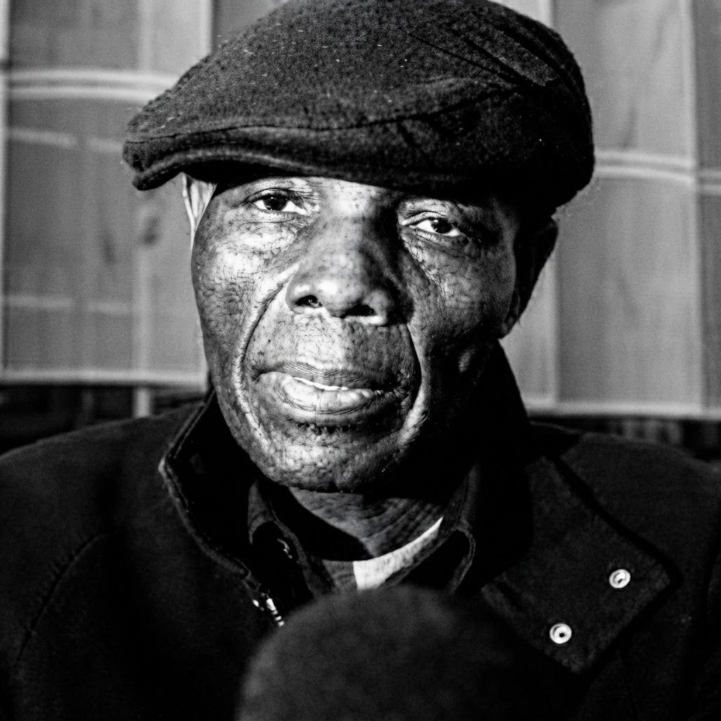 Tuku in his flat cap, microphone in hand — black and white portrait