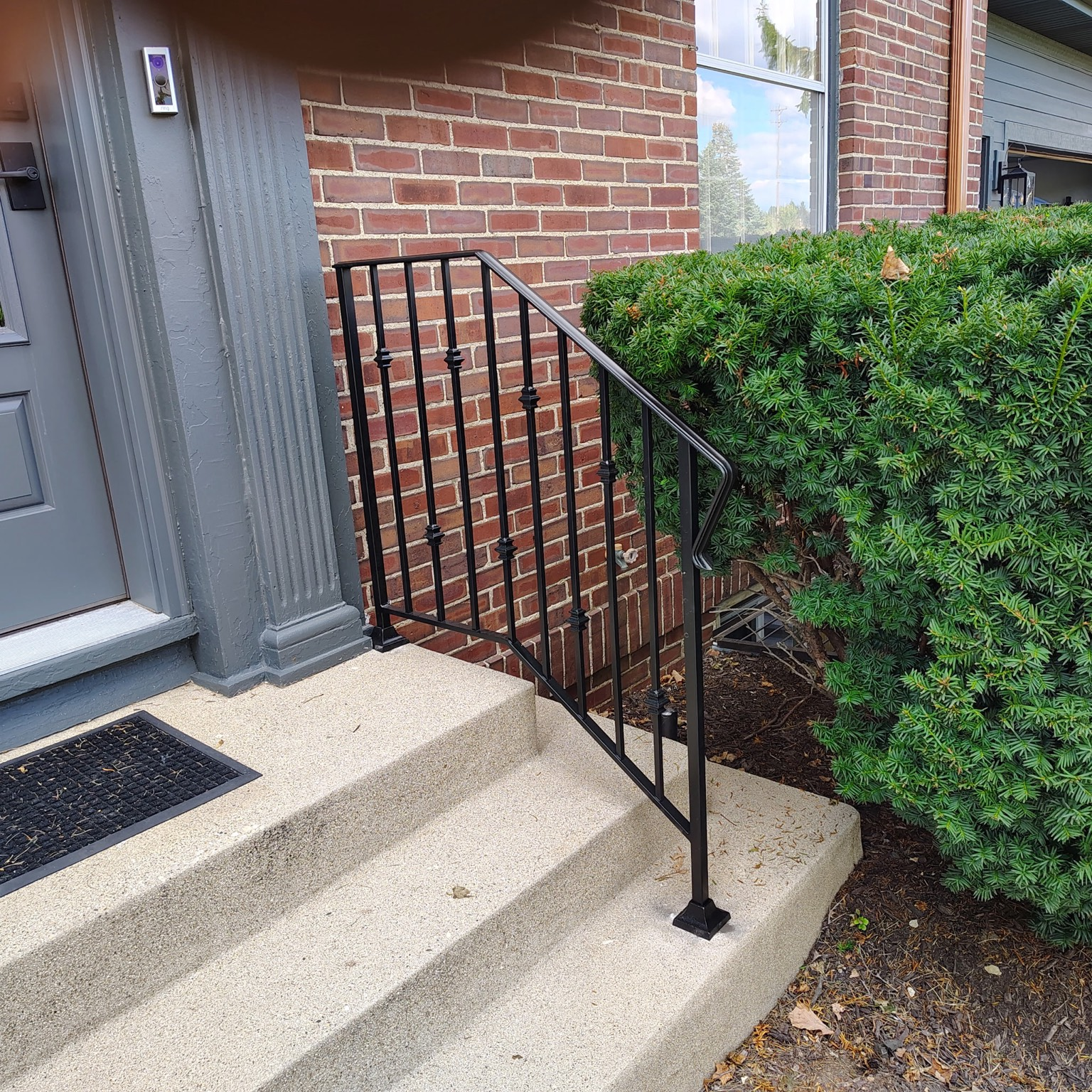 Black metal handrail with vertical balusters on a concrete step at a brick residence.
