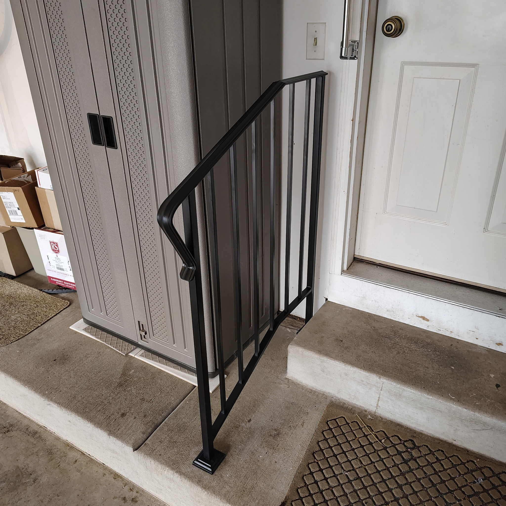 Black metal handrail with vertical balusters bolted to a two-step concrete entry.