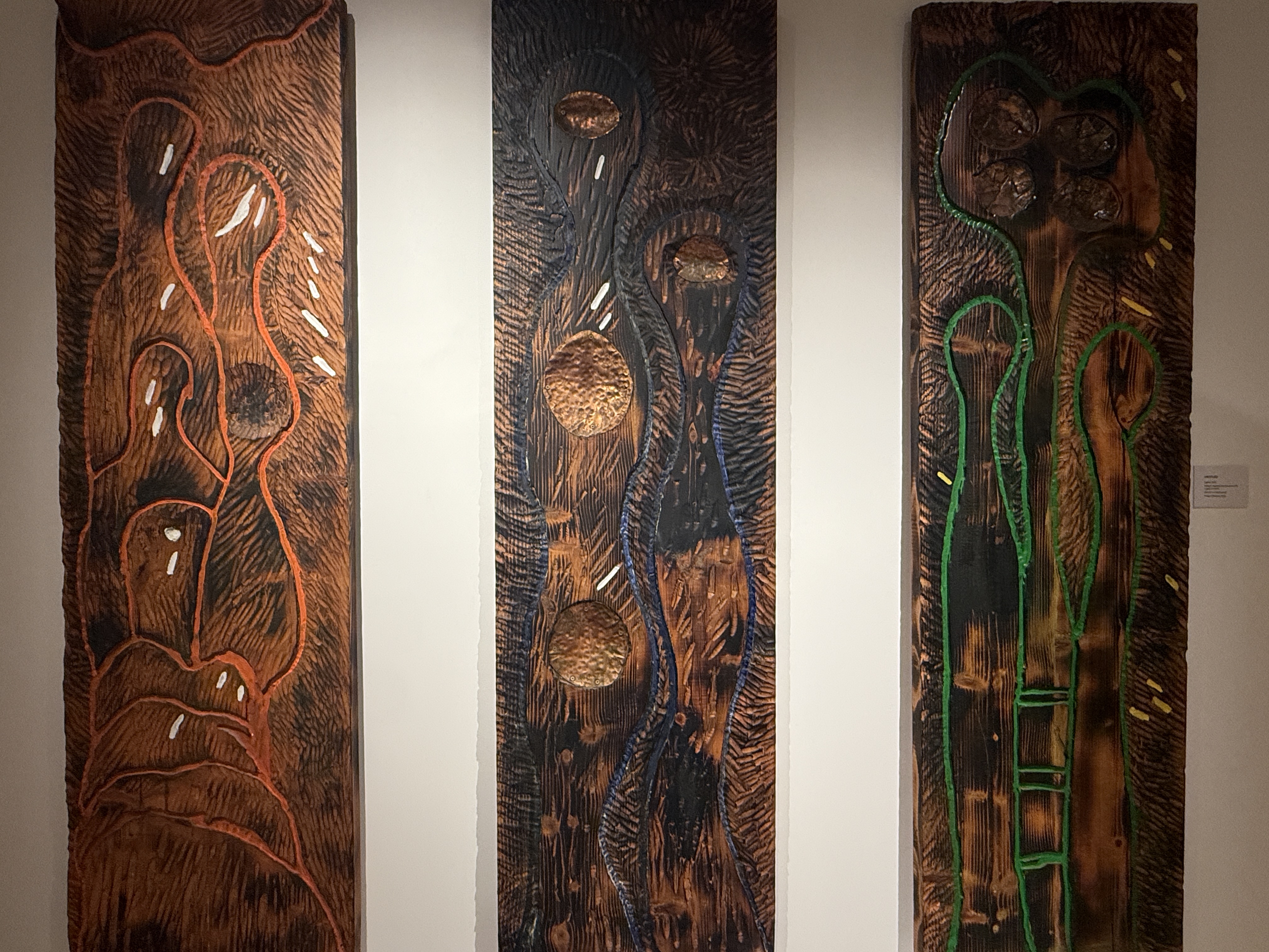Three vertical carved wood relief panels by Gabriel Caruana with copper discs, orange and green pigments at the Victor Passmore Gallery Valletta