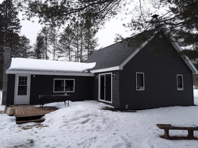 Rear exterior of a single-story home with dark grey horizontal siding, white trim, a wooden deck and a stone chimney