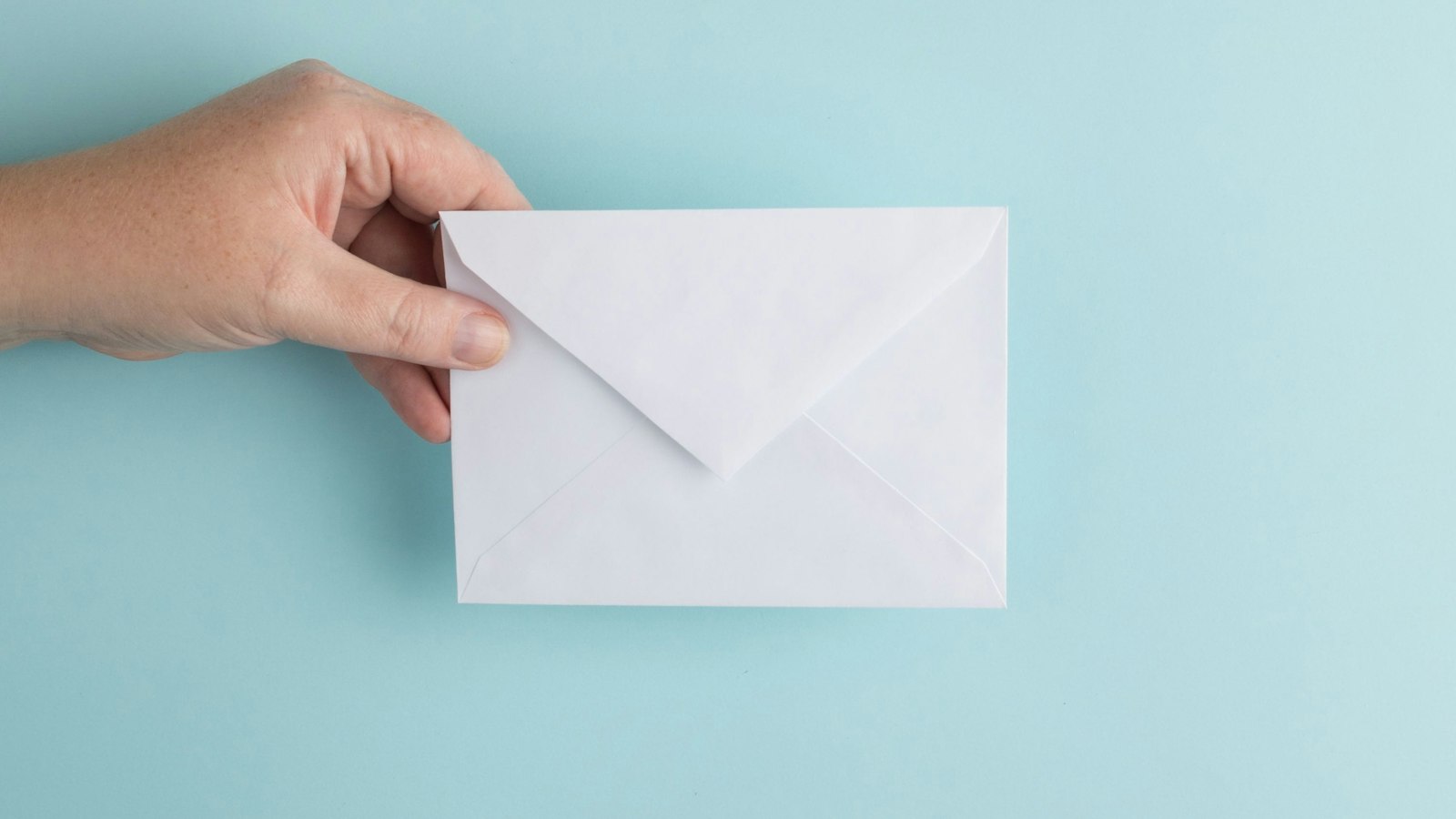 A hand holding up a plain white sealed envelope against a pale-blue background