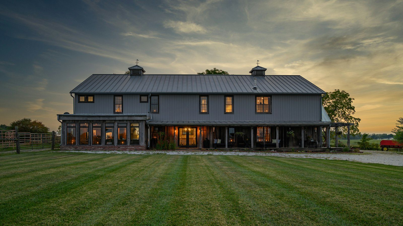 Large dark-gray farmhouse-style home with lit windows and a wide lawn at sunset