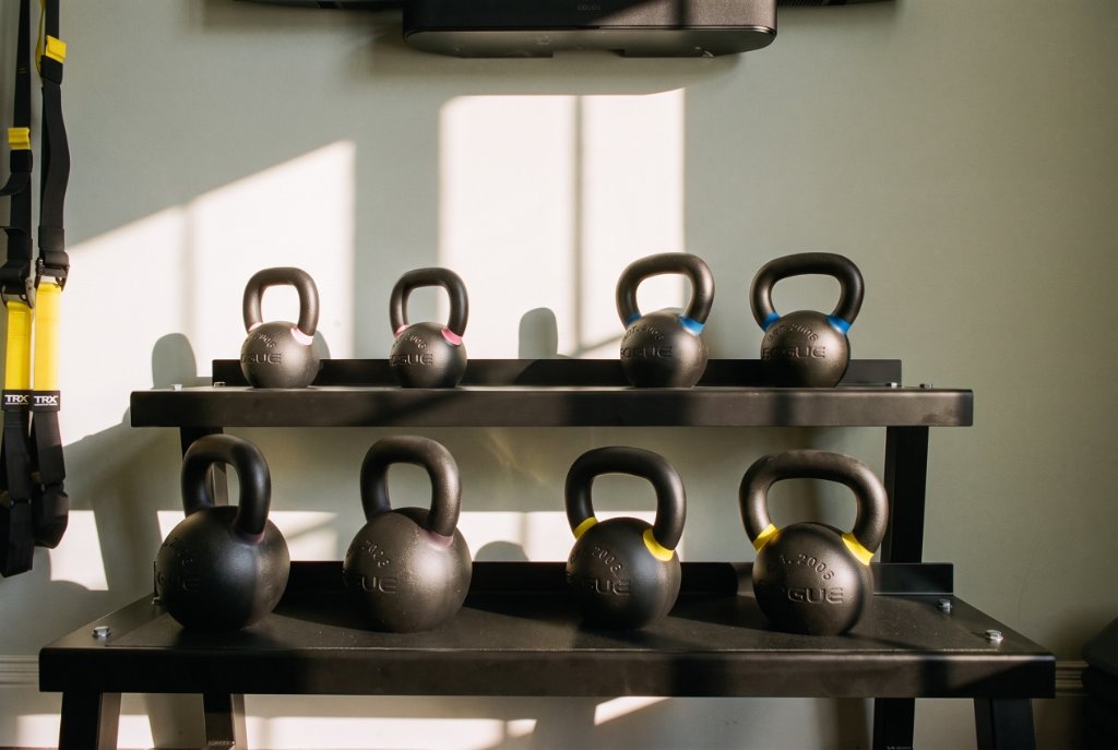Two-tier black metal kettlebell rack with Rogue kettlebells, TRX suspension trainer and wall-mounted soundbar in the Oak & Iron gym