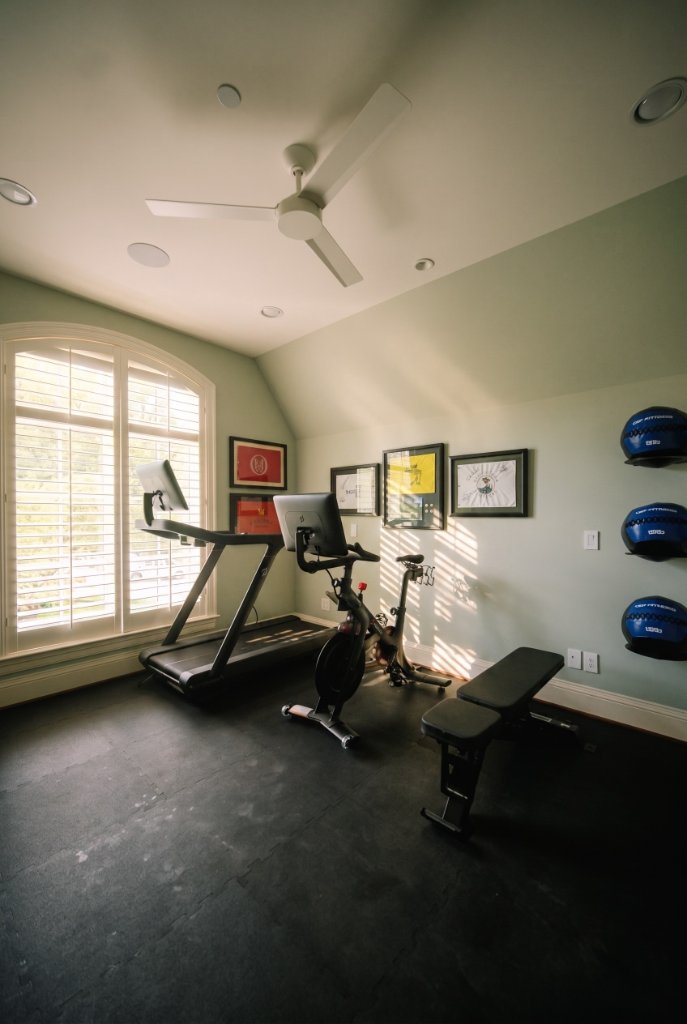 Corner view of the Oak & Iron home gym with arched window, treadmill, exercise bike, weight bench and wall-mounted medicine balls