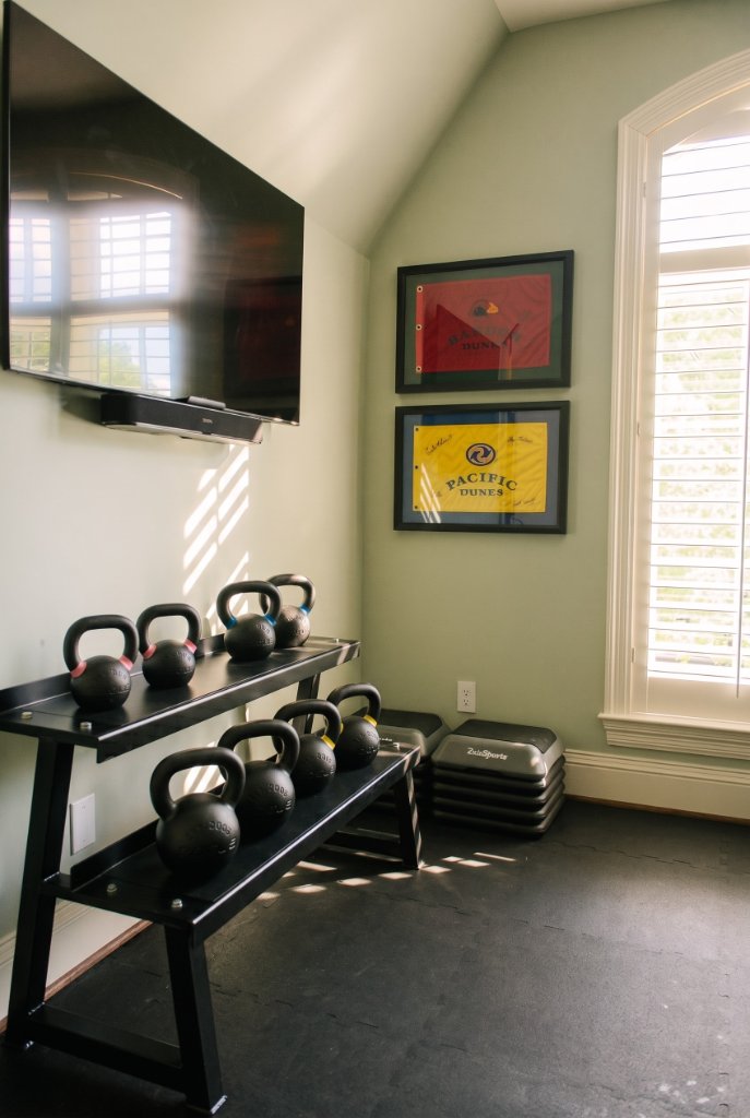Interior view of the Oak & Iron home gym showing kettlebell rack, wall-mounted TV, treadmill and exercise bike with natural light from the arched window