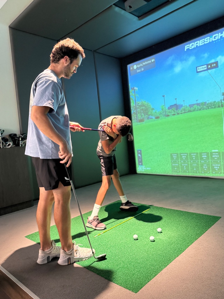 Coach guiding a junior student through a swing on a Foresight Sports indoor simulator
