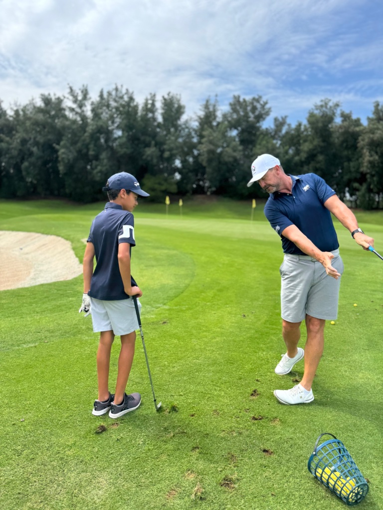 Coach instructing a junior golfer outdoors on a sunny golf course fairway