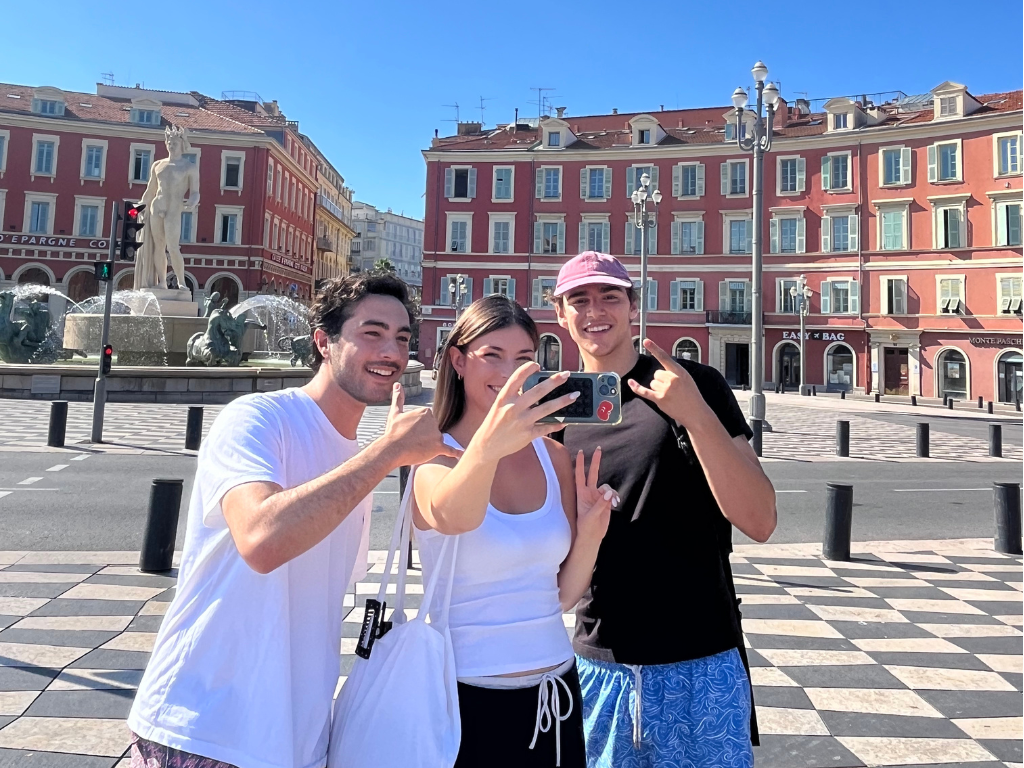 Trois amis prenant un selfie sur une place historique de Nice avec une fontaine classique et des bâtiments ocre en arrière-plan