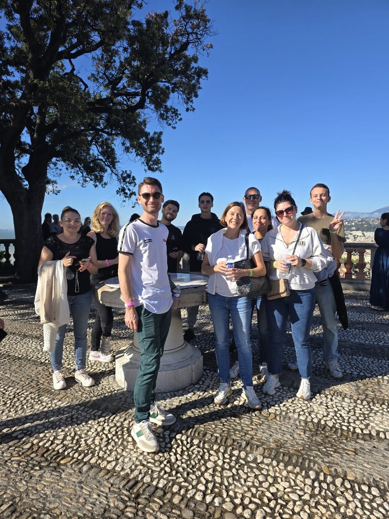 Grand groupe posant autour d'un repère en pierre sur une place de Nice avec ciel bleu