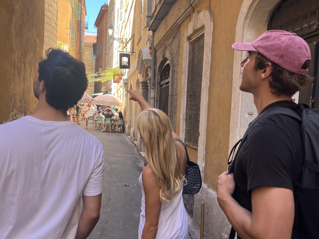 Trois amis marchant dans une rue étroite et ensoleillée de Nice, l'une pointant vers un café