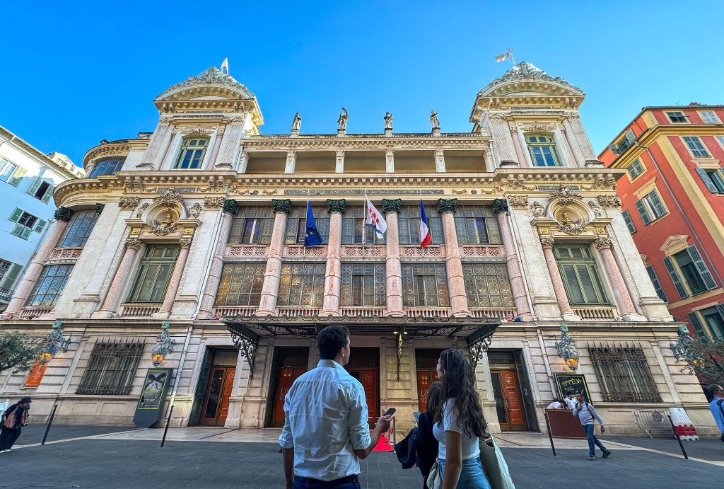 Deux visiteurs contemplant la façade ornée de l'Opéra de Nice sous un ciel bleu