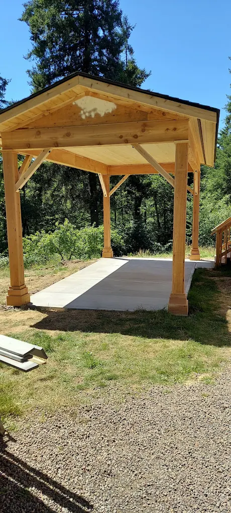 Timber-frame open pavilion with decorative post bases, knee braces, and a fresh concrete slab floor.