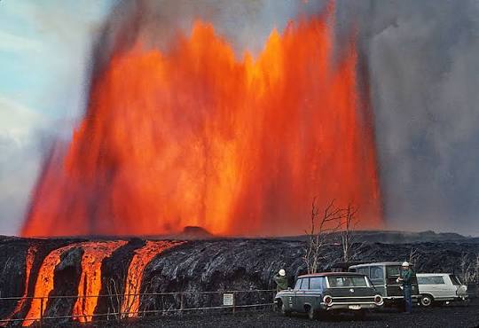 Historic photograph of a massive volcanic eruption with a fountain of molten lava, billowing ash, and observers parked nearby on a black lava field.