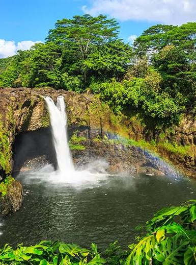 Vertical photograph of Akaka Falls plunging into a dark pool surrounded by lush tropical rainforest, with a rainbow visible through the mist.