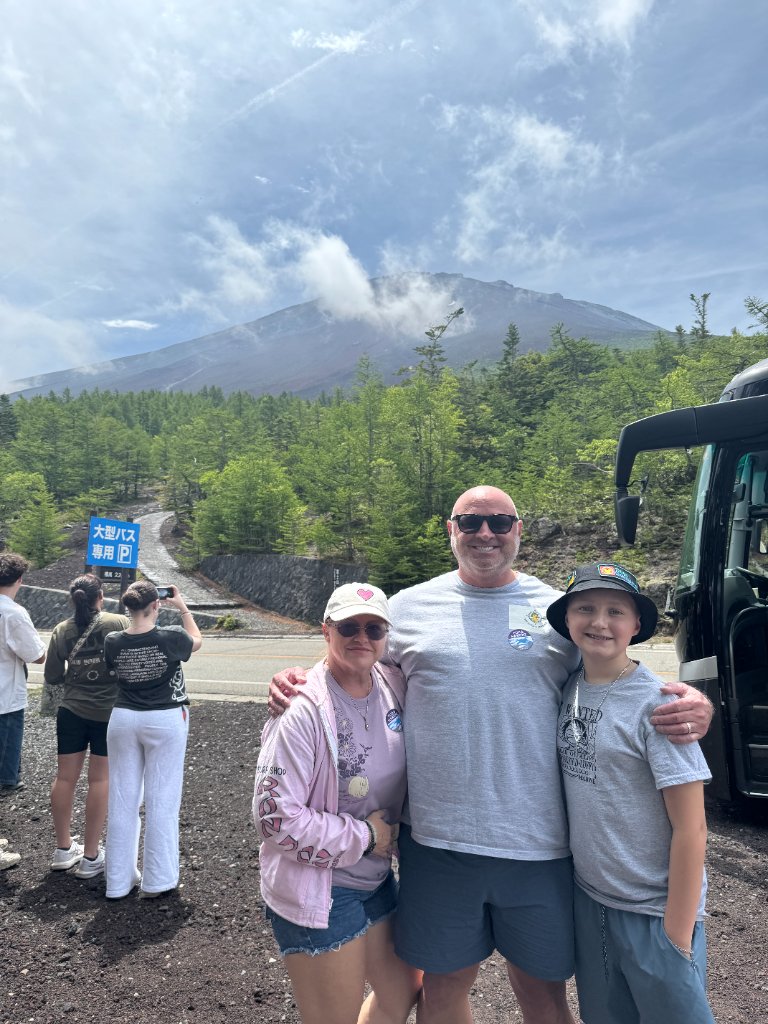 A family of three posing together at the base of Mount Fuji under a blue sky with scattered clouds