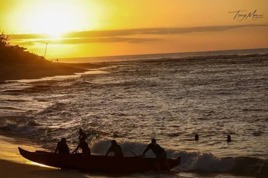 Photograph of a traditional Hawaiian outrigger canoe being launched into the surf at sunset, with crew silhouetted against a brilliant orange horizon.