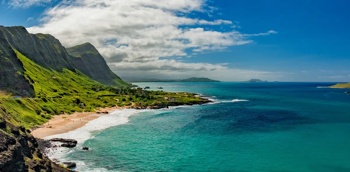 Wide panoramic photograph of a Hawaiian coastline with steep green volcanic cliffs, a golden beach, and turquoise ocean transitioning to deep sapphire blue.