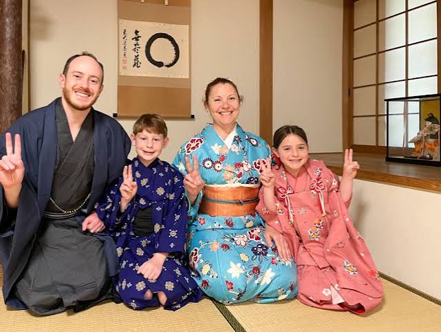 A family of four in traditional yukatas and kimonos kneeling on tatami mats, all making peace signs