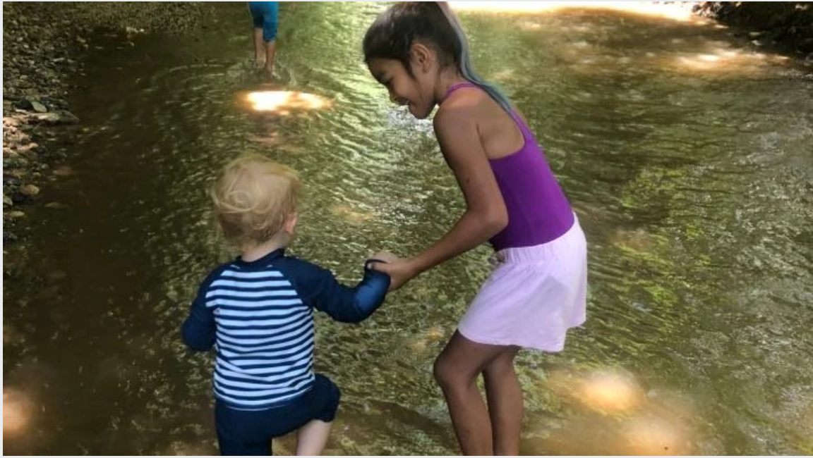 An older child holding a toddler's hand as they wade together in a shallow sunlit stream