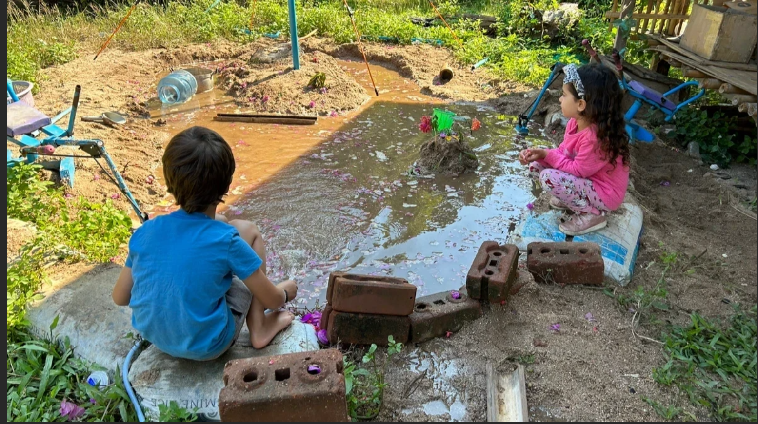 Two children playing at a muddy pool bordered by red bricks