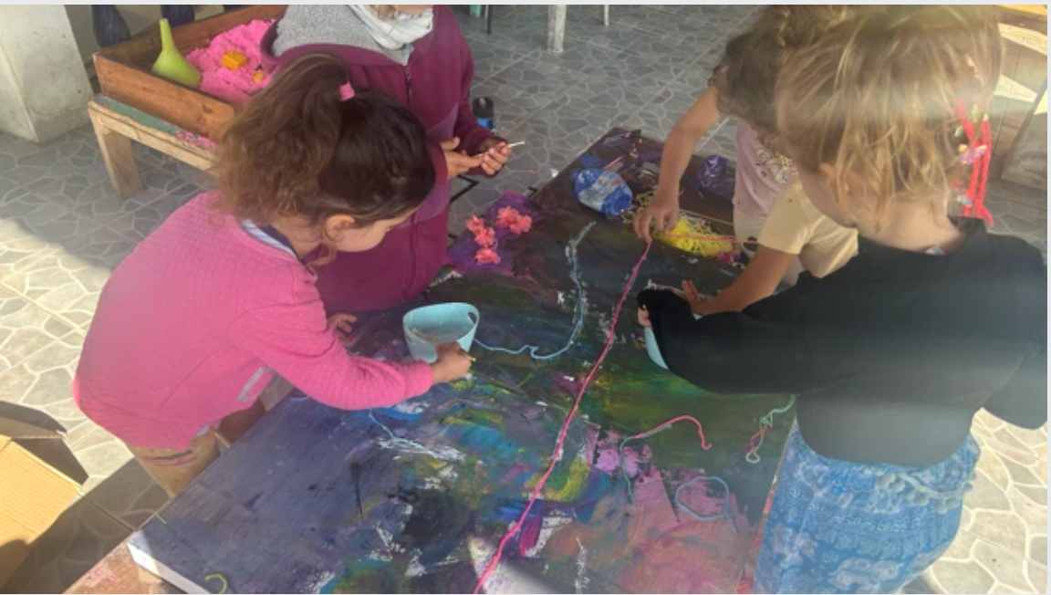 Children gathered around a table covered in dark paint, yarn, and pink flowers during a collaborative art project