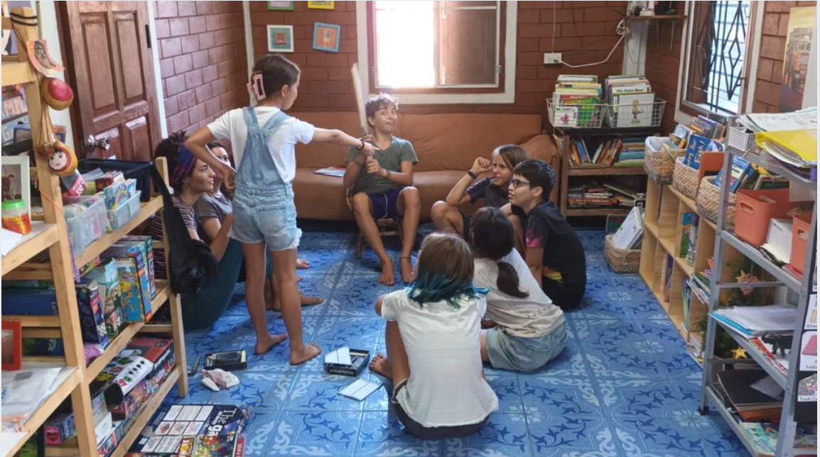 Children and facilitators gathered in a circle on the blue-tiled floor of the unschooling community room
