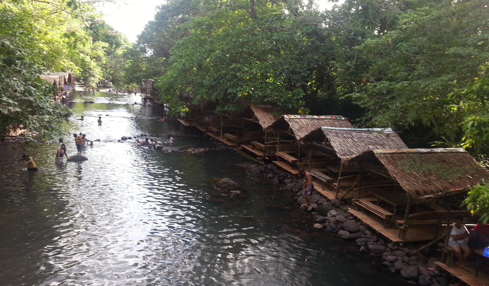 Panguil River Eco-Park: bamboo huts along the riverbank under dense rainforest canopy, with visitors wading in the shallows.
