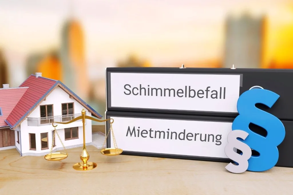 Zwei schwarze Aktenordner mit Aufschriften „Schimmelbefall" und „Mietminderung" neben goldener Justitia-Waage, Paragraphen-Symbolen und einem Hausmodell
