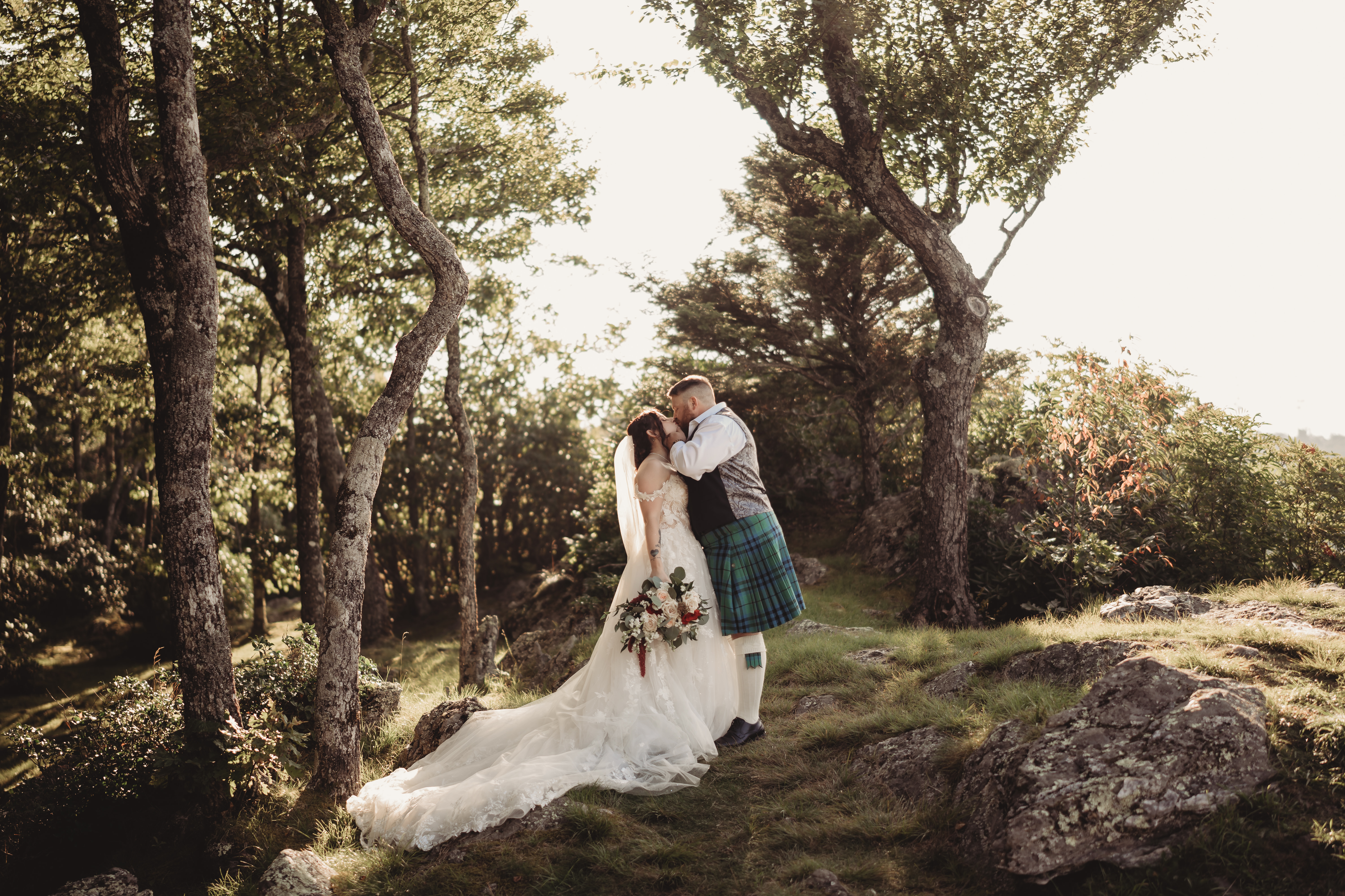 A bride and groom share a kiss in a sunlit forest, her lace gown and train pooling on the grass beside his kilt and waistcoat.