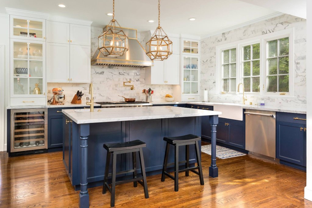 San Mateo kitchen with navy-blue shaker cabinetry, brushed-gold hardware, a full-height marble backsplash and island, farmhouse sink, and gold geometric pendants.