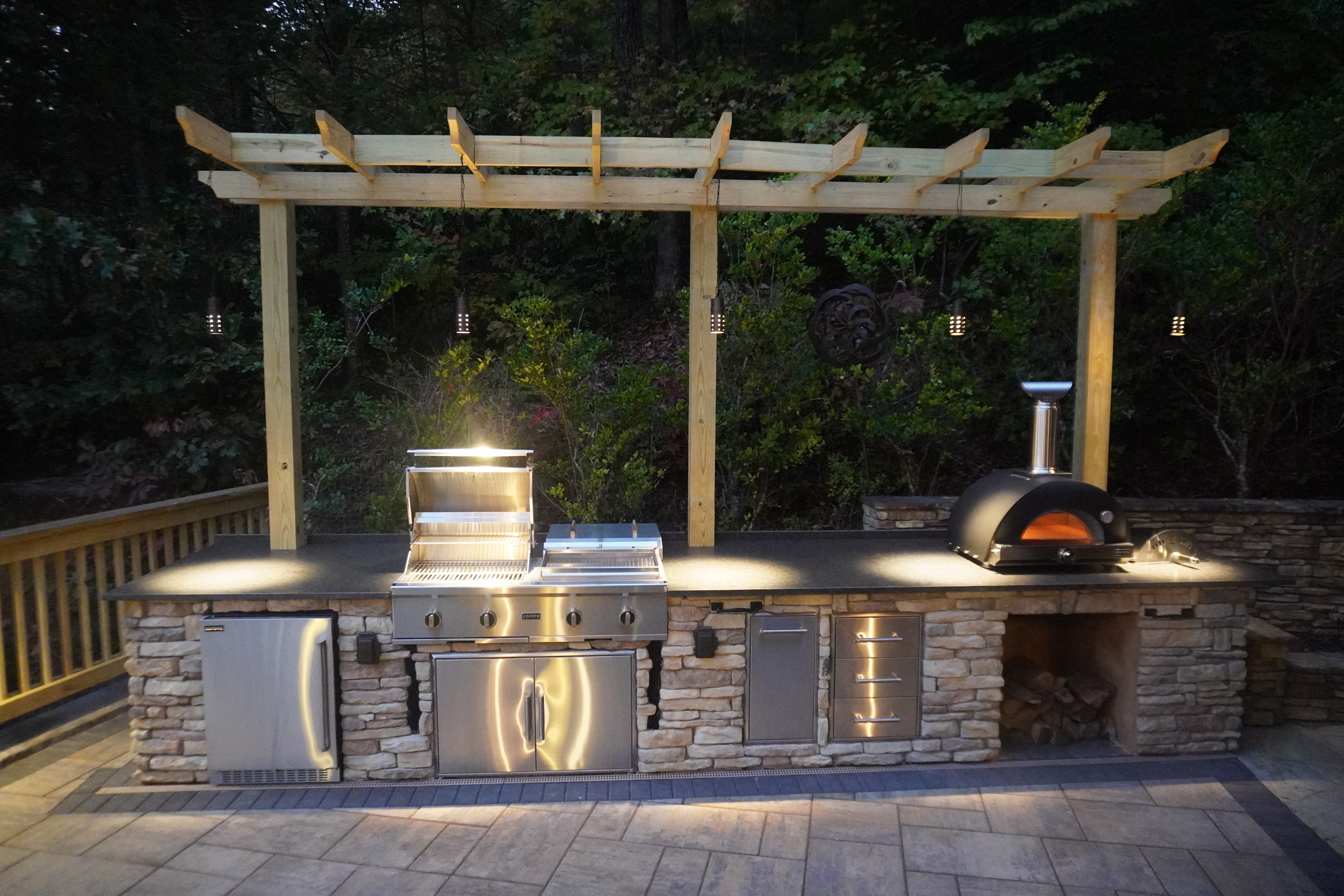 Custom stone outdoor kitchen with built-in grill, pizza oven and wood pergola with pendant lighting in Knoxville TN by Grateful Grounds