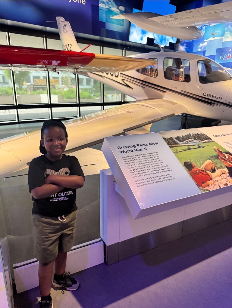 Cirrus Airplane Display Visit Child standing confidently in front of a Cirrus airplane display during a STEM museum field trip organized by Insight Kidz Care