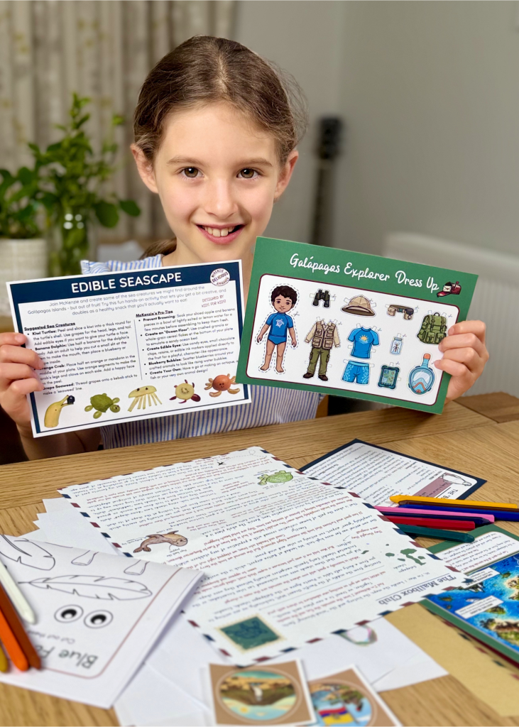 A smiling girl holding her Mailbox Club activity sheets — the Edible Seascape recipe and Galápagos Explorer Dress Up — above a table spread with her adventure letter, postcards, stickers and colouring pencils