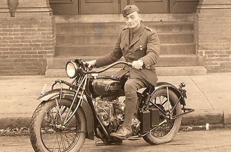 Sepia vintage photo of a man in military uniform on an early Indian motorcycle