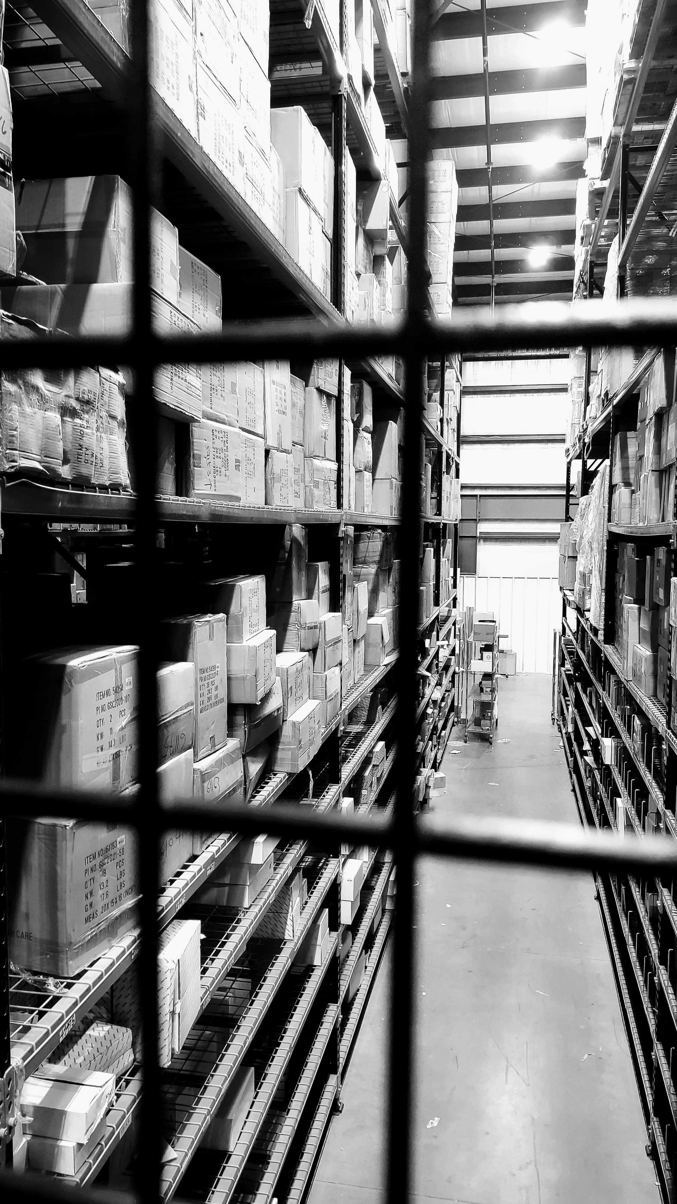 Black and white photograph looking down a warehouse aisle stacked with cardboard boxes