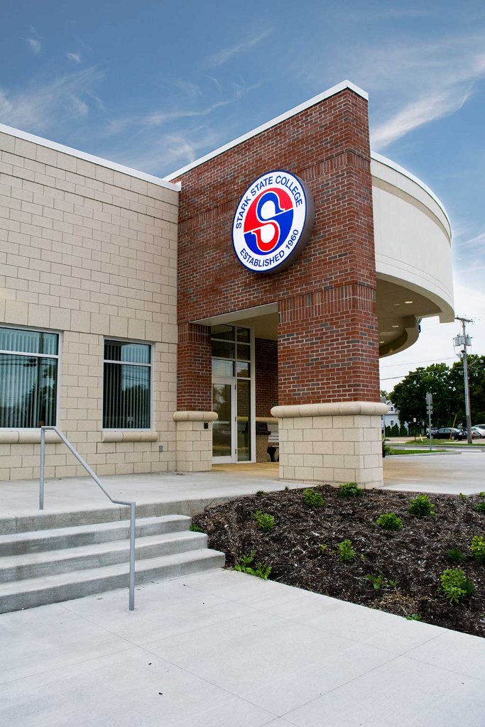 Night exterior of Stark State College Alliance Satellite Center in Alliance, OH — brick and masonry civic building with illuminated entry canopy, large glazing, and branded signage glowing against a deep blue sky